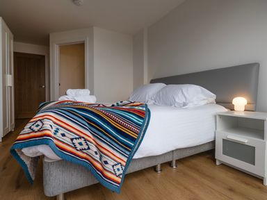 A bedroom with a bed covered with white sheets and a colorful blanket with a bedside table and lamp at Treen in St Ives