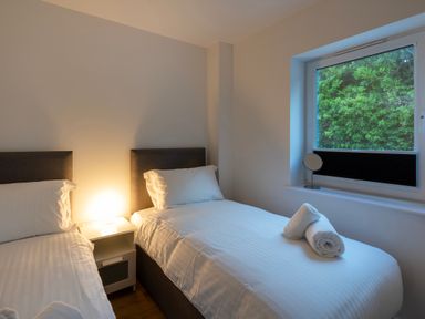 A bedroom with two single beds a bedside table with a lamp and a window showing green foliage at Treen in St Ives
