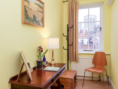 A small room with a wooden desk holding a lamp plant and notebook a wooden chair near a window with a view of rooftops at 6 Granary Court in York