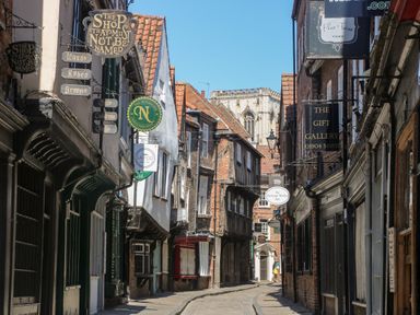A narrow cobblestone street with shops and hanging signs in York with a church visible in the background at 6 Granary Court York