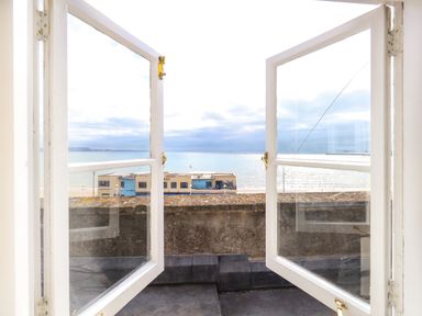 An open window overlooking a balcony wall with a beach building and the sea at The Esplanade in Weymouth