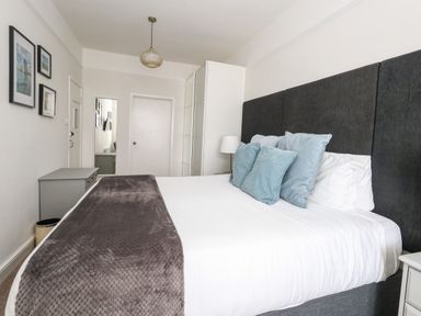 A bedroom with a bed with a dark headboard and blue pillows a grey blanket a bedside lamp a dresser and framed pictures on the wall at The Esplanade in Weymouth