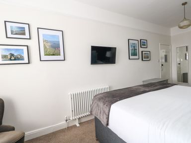 A bedroom with a bed with white sheets and gray blanket a wall-mounted TV framed pictures a radiator a chair and a mirror at The Esplanade in Weymouth