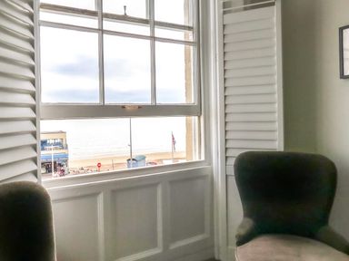 Two chairs near a window with shutters overlooking a beach and buildings at The Esplanade in Weymouth