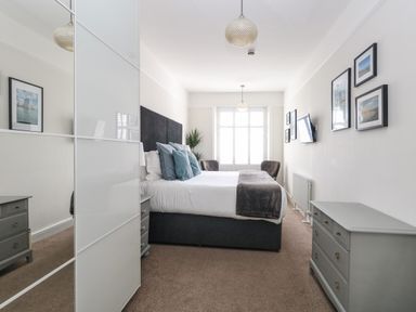 A bedroom with a bed with pillows and a blanket two chairs near a window a TV on the wall and dressers at The Esplanade in Weymouth