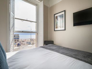 A bedroom with a bed a wall-mounted TV a framed picture and a window with shutters overlooking a street and sea at The Esplanade in Weymouth