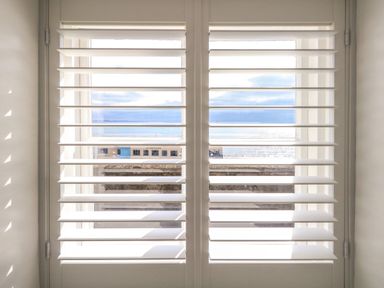 A window with white shutters partly open showing a sea view at The Esplanade in Weymouth