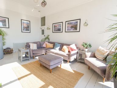 A living room with a gray sectional sofa an ottoman side tables framed pictures and plants at The Esplanade in Weymouth