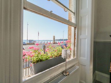 A window with pink flowers in a planter and a lamp on a radiator below overlooking a street and sea at The Esplanade in Weymouth