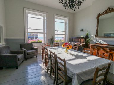 A dining room with a table and chairs a vase of flowers two armchairs a piano and a large mirror at The Esplanade in Weymouth