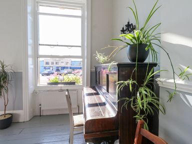 A room with a wooden upright piano and plants near a large window at The Esplanade in Weymouth