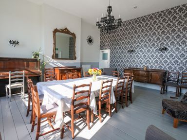 A dining room with a long table covered by a white tablecloth surrounded by wooden chairs a piano a large mirror a chandelier and patterned wallpaper at The Esplanade in Weymouth