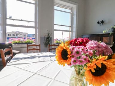 A dining room with a table covered by a white cloth and a vase of flowers near large windows with a view of a building at The Esplanade in Weymouth