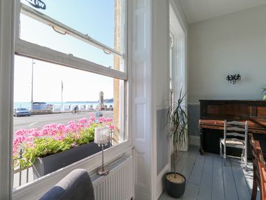 A room with a piano a chair a potted plant and a window showing pink flowers and a view of the sea at The Esplanade in Weymouth