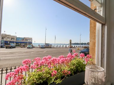 A view of a street with cars and a building across the road with the sea and a promenade in the background with pink flowers on a window ledge at The Esplanade in Weymouth
