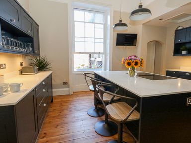 A kitchen with black cabinets wooden floor stools around island and flowers on the counter at The Esplanade in Weymouth