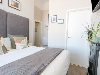 A bedroom with a bed with cushions and a gray headboard a door a fireplace with plants and pictures on the wall at The Esplanade in Weymouth