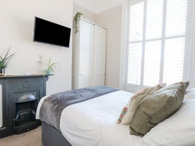 A bedroom with a bed with pillows a wall mounted tv a fireplace plants a wardrobe and large windows with shutters at The Esplanade in Weymouth