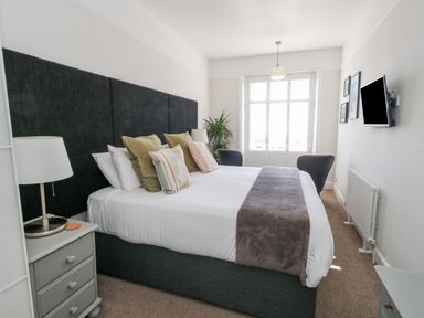 A bedroom with a large bed with multiple pillows bedside tables lamps a wall-mounted TV and two chairs at The Esplanade in Weymouth