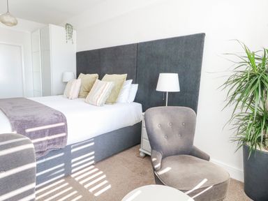 A bedroom with a large bed with pillows a gray chair a white nightstand with lamp a wardrobe and a potted plant at The Esplanade in Weymouth