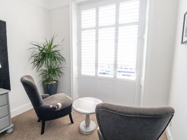 A small sitting area with two gray chairs, a white round side table, a potted plant, and window shutters at The Esplanade in Weymouth