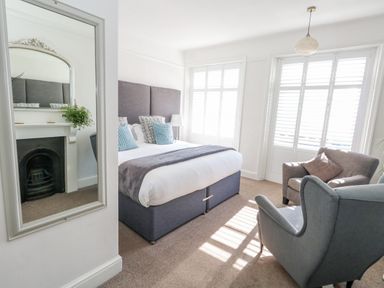 A bedroom with a bed, two armchairs, a large mirror, a fireplace, and windows with shutters at The Esplanade in Weymouth