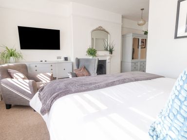 A bedroom with a bed a gray blanket two armchairs a TV on the wall a dresser a mirror above a fireplace and a wardrobe at The Esplanade in Weymouth