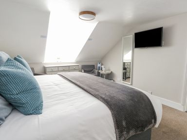 A bedroom with a bed with blue pillows and a gray blanket a dresser a small table and chair a wall-mounted TV and a full-length mirror at The Esplanade in Weymouth