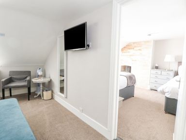 A sitting area with a gray armchair a small round table a wall-mounted TV and a view into a bedroom with two beds and a chest of drawers at The Esplanade in Weymouth
