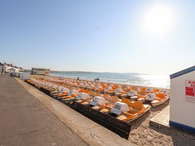 A row of orange and white pedal boats on a beach near the sea with a paved walkway on the left at The Esplanade in Weymouth