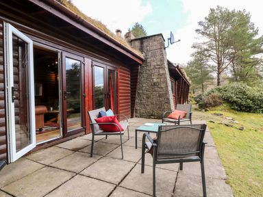 An outdoor patio with three chairs and a table beside a wooden cabin with open glass doors and trees in the background at Rocksite in Kirkmichael Perth And Kinross