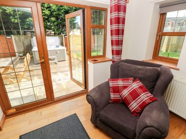 A living area with a gray armchair and red plaid cushions near a glass door leading to a patio with outdoor furniture and a jacuzzi at Ty Melyn in Boncath