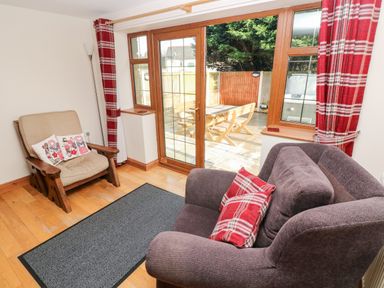 A living room with armchairs red plaid cushions and curtains and glass doors leading to a patio at Ty Melyn in Boncath