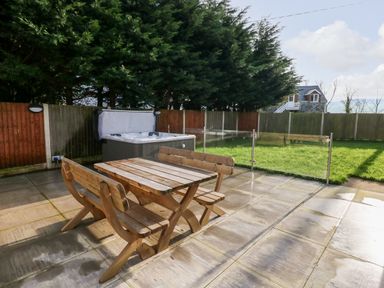 A garden patio with a wooden picnic table and benches a hot tub and a glass fence enclosing a grassy area at Ty Melyn in Boncath