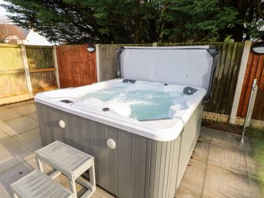 An outdoor hot tub with bubbling water and a gray cover partially open on a tiled patio with wooden steps and fenced yard at Ty Melyn in Boncath