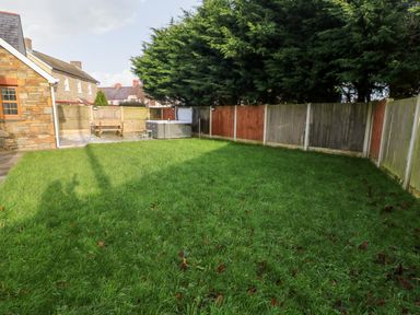 A backyard with a grassy lawn a tiled patio wooden bench and a hot tub surrounded by a wooden fence and trees at Ty Melyn in Boncath