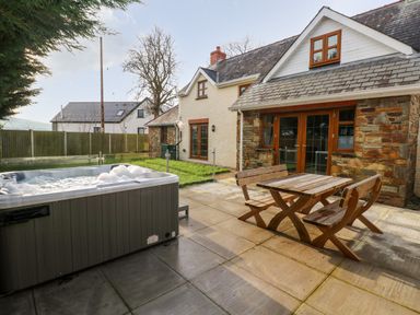 A patio with a wooden table and benches next to a hot tub outside a stone and brick house at Ty Melyn in Boncath