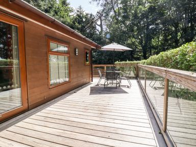 A wooden deck with table chairs and an umbrella next to a building with windows surrounded by trees and greenery at Swinsty Lodge in Fewston near Darley