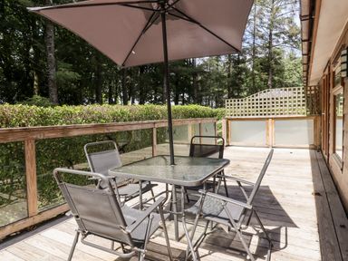 An outdoor wooden deck with a glass table and four metal chairs under a large umbrella surrounded by trees at Swinsty Lodge in Fewston near Darley