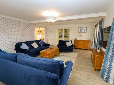A living room with blue sofas a wooden coffee table wooden cabinets patterned curtains and double glass doors at Drifters in Winterton-On-Sea