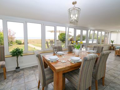A dining room with a wooden table set with plates and glasses surrounded by striped chairs by large windows at Drifters in Winterton-On-Sea