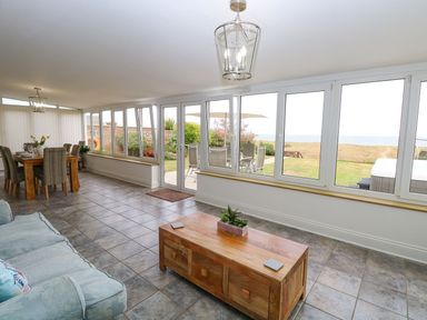 A room with large windows overlooking a garden and sea with a wooden coffee table a sofa and a dining table at Drifters in Winterton-On-Sea