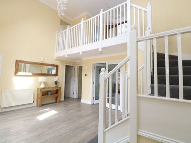 An entrance hall with light wood flooring a wooden console table with a lamp and flowers a large wall mirror white banisters and stairs at Drifters Winterton-On-Sea