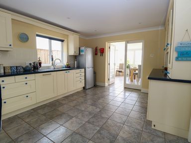A kitchen with cream cabinets and blue knobs a stainless steel fridge a tiled floor and a dining area through glass doors at Drifters in Winterton-On-Sea