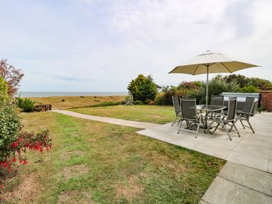 A garden with a patio table and chairs under an umbrella and a path leading to the sea at Drifters in Winterton-On-Sea