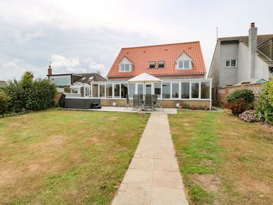 A backyard with a paved path leading to a patio with outdoor dining set and umbrella beside a hot tub a two-story house with red tile roof and large windows at Drifters in Winterton-On-Sea