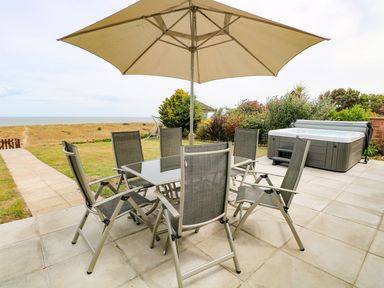 A patio with a dining table and chairs under an umbrella and a hot tub overlooking grass and beach at Drifters in Winterton-On-Sea