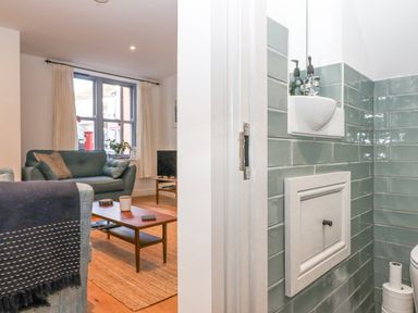 A living room with a blue sofa and wooden coffee table next to a bathroom with green tiles and a small white sink at The Cottage on The Square in Chagford