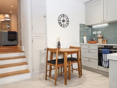 A kitchen with a wooden table and two chairs a clock on the wall and stairs leading to a living area at The Cottage on The Square in Chagford