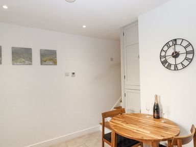 A dining area with a round wooden table and two chairs a bottle and two glasses on the table a wall clock and three square pictures on the wall at The Cottage on The Square in Chagford